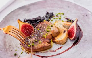 Foie-gras-figues-scaled Close-up of a plated gourmet dish: sliced seared meat with figs, crushed pistachios, and a berry-like sauce on a pale plate