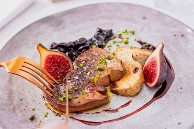 Foie-gras-figues-scaled Close-up of a plated gourmet dish: sliced seared meat with figs, crushed pistachios, and a berry-like sauce on a pale plate